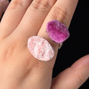 Puede incluir: Dos anillos con piedras en bruto sin pulir. Un anillo presenta una piedra ovalada de color rosa suave, mientras que el otro anillo tiene una piedra de color magenta intenso con una superficie cristalina texturizada sobre una banda metálica.