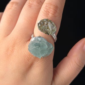 Könnte beinhalten: Ein offener Ring mit zwei verschiedenen Rohsteinen. Ein Stein ist ein durchscheinender, blass minzgrüner Kristall mit facettiertem Aussehen, während der andere ein kleinerer, texturierter, metallisch goldener und grauer Mineralcluster auf einem Silberring ist.