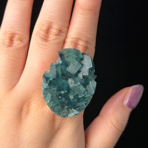 Op de afbeelding: Een opvallende ring met een grote, ruwe cluster van blauwgroene en talingkleurige fluorietkristallen. Het mineraal vertoont een natuurlijke, grillige formatie met meerdere kubusvormige facetten en doorschijnende randen op een metalen ringband.