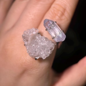 May include: A silver-toned ring featuring two natural crystals. One crystal is a cluster of small, clear points, and the other is a larger, elongated, pale purple crystal. The ring is set against a blurred background.