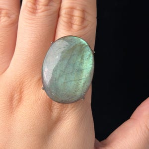Può includere: Un grande anello con pietra di labradorite di forma ovale, caratterizzato da una superficie liscia e lucida con riflessi iridescenti verdi e blu, montato su una semplice struttura in metallo con griffe che fissano la pietra.