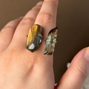 Puede incluir: Un anillo de doble piedra que presenta una labradorita ovalada con iridiscencia marrón dorada y un cristal de cuarzo transparente, fino y alargado. Ambas piedras están montadas en una banda metálica que rodea el dedo con un diseño abierto.