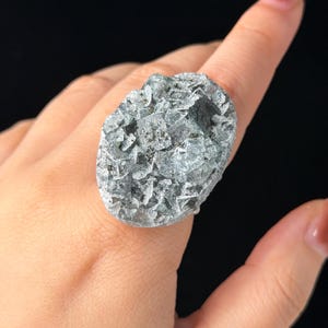 Könnte beinhalten: Ein Statement-Ring mit einer großen, ovalen Ansammlung roher, kristalliner Mineralformationen. Die Oberfläche des Steins zeigt ein strukturiertes, metallisch silbergraues Aussehen mit facettierten Kanten und tiefen, zerklüfteten Spalten.
