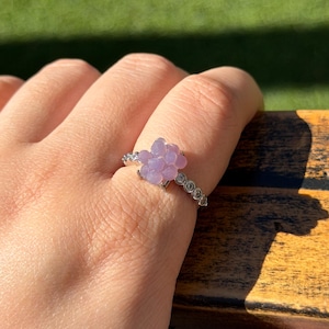 May include: A silver ring with a cluster of light purple, flower-shaped beads. The band is decorated with small, clear stones. The ring is displayed on a finger, with a blurred green background.