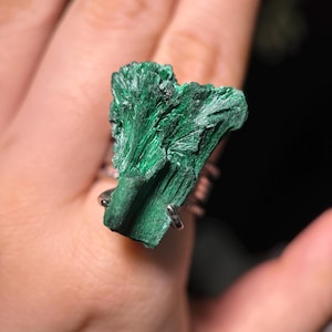 Puede incluir: Anillo de plata con una piedra de malaquita verde vibrante y texturizada. La malaquita tiene una apariencia cristalina y en capas, con diferentes tonos de verde. La banda del anillo es delgada y parece ser de plata.