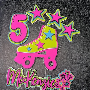 May include: A neon pink and green roller skate cake topper with the number 5, stars, and the name "McKenzie". The design features a retro color scheme with a black glitter outline, perfect for a birthday celebration.
