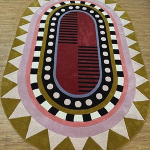 May include: An oval-shaped rug with a geometric design. The rug features a central maroon rectangle with black stripes, surrounded by concentric rings of black, white, pink, and olive green. White triangles border the outer edge.