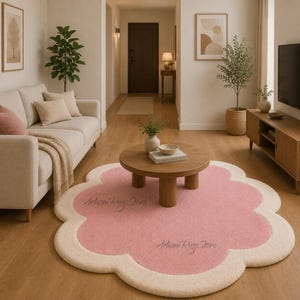 Op de afbeelding: Een roze en crèmekleurig, wolk-vormig tapijt ligt op een houten vloer in een woonkamer. Het tapijt heeft een zachte, pluche textuur en draagt de tekst "Arham Rugs Store". Een ronde houten salontafel staat in het midden.