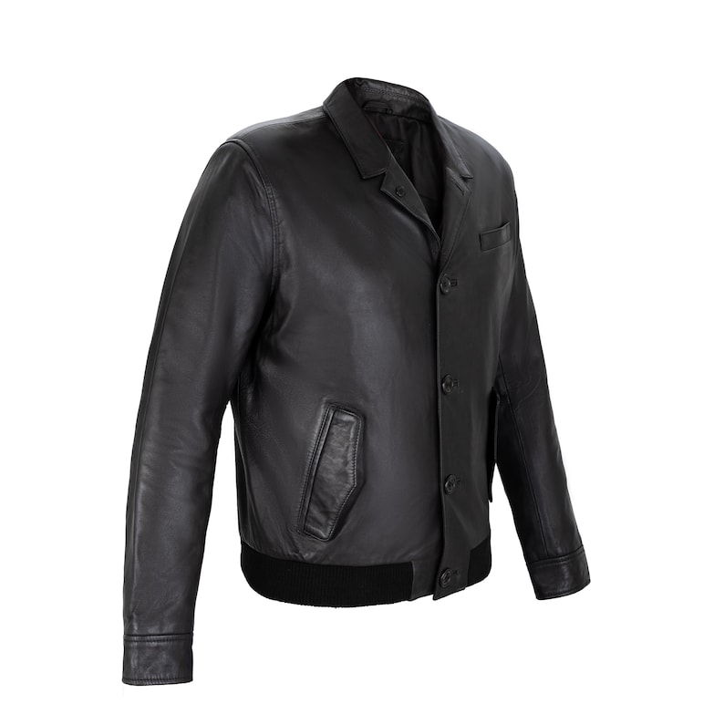 May include: A black leather jacket featuring a pointed collar, a front button closure with four visible buttons, two slanted side pockets, and a ribbed black hem. The jacket has long sleeves with buttoned cuffs and a smooth, matte leather finish.