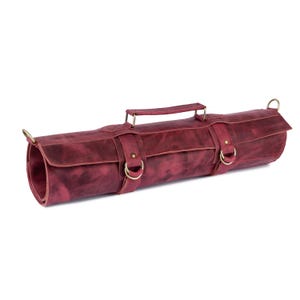 Könnte beinhalten: Eine aufgerollte, weinrote Leder-Messertasche mit Griff und messingfarbener Hardware. Die Rolle hat zwei Riemen mit Schnallen und Messingringen an jedem Ende. Das Leder hat ein abgenutztes Aussehen.