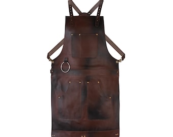 May include: A brown leather apron with adjustable straps and multiple pockets. The apron features a large front pocket, two smaller pockets, and a metal ring. The leather has a distressed appearance with dark shading.