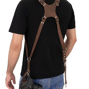 May include: A brown leather camera harness with adjustable straps and metal fixings. The harness is designed to distribute the weight of a camera, as seen attached to a DSLR camera with a lens. The harness is worn over the shoulders and across the back.