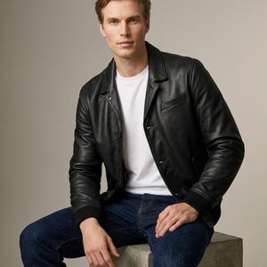 May include: A black leather jacket with a collared neckline, long sleeves with ribbed cuffs, and a button-front closure, worn over a plain white t-shirt and dark blue denim jeans. The jacket features a smooth texture and a structured fit.