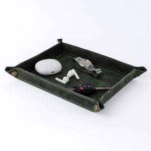 May include: A dark green leather tray containing a watch, car keys, wireless earbuds, and a charging case. The tray has a stitched border and brass-coloured snap closures. The items inside are white and silver.