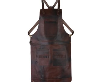 May include: A brown leather apron with adjustable straps and multiple pockets. The apron has a chest pocket, two large front pockets, and smaller pockets for tools. The apron is designed for use in a workshop or kitchen.