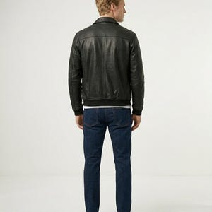 May include: A black leather jacket with a classic collar, long sleeves, and a ribbed hem, paired with dark blue denim jeans. The jacket features a smooth texture and visible vertical seam detailing on the back, worn over a plain white shirt.