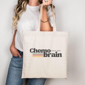 May include: Cream-coloured tote bag featuring the text "Chemo brain since 2024" in dark grey. Below the text is a horizontal stripe design in orange, green, and yellow. The bag has long handles.