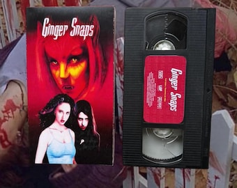 May include: A VHS tape and its box for the film "Ginger Snaps." The box has a red background with a woman's face and two other women. The VHS tape is black with a red label that also says "Ginger Snaps."