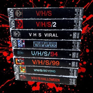 Op de afbeelding: Een stapel zwarte VHS-banden met titels als "V/H/S", "V/H/S/2" en "V/H/S/85" in rode en witte letters. De banden hebben een "VHS"-logo en "hi-fi" en "Dolby Digital" labels. De achtergrond is zwart met rode spetters.