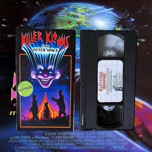 Puede incluir: Caja y cinta VHS de la película de terror "Killer Klowns from Outer Space". La portada presenta una cara de payaso amenazante sobre un fondo estrellado, con la palabra "HORROR". La cinta VHS es negra con una etiqueta blanca.
