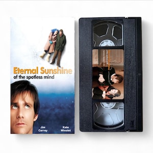 Puede incluir: Una cinta VHS y su carátula de la película "Eternal Sunshine of the Spotless Mind". La carátula presenta a Jim Carrey y una pareja con ropa de invierno. La cinta VHS negra tiene una etiqueta con una escena de la película.