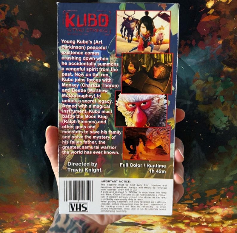 May include: A VHS tape for the animated film "Kubo and the Two Strings." The cover features colorful illustrations of characters and scenes from the movie, with text describing the plot and cast. The tape is held in a hand.