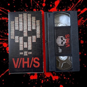 Può includere: Una videocassetta VHS nera e la sua custodia. La custodia presenta un design a teschio pixelato e il testo "V/H/S" in rosso. La videocassetta VHS ha anche il testo "V/H/S" in rosso, insieme ad altro testo.