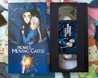 May include: A VHS tape and its cover for the animated film "Howl's Moving Castle." The cover features a blue and black illustration of two characters with the film's title in white. The VHS tape is black with a label that matches the cover art.