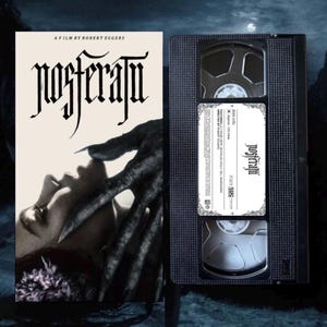 Puede incluir: Una cinta VHS y un póster de película vintage de la película "Nosferatu". El póster presenta el rostro de una mujer con una mano oscura que se extiende hacia ella. La cinta VHS tiene el título de la película en su etiqueta. El texto "A FILM BY ROBERT EGGERS" es visible.