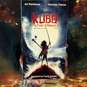 May include: A movie poster for "Kubo and the Two Strings" featuring Art Parkinson and Charlize Theron. The poster shows a silhouetted figure holding a sword and a musical instrument, standing on a small island in front of a large wave and a full moon.