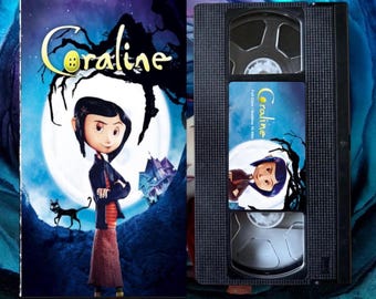May include: A VHS tape of the animated film "Coraline." The cover art shows a girl in a black jacket and a long skirt, standing before a full moon. A black cat is to her left, and a spooky house is in the background.