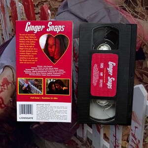 Puede incluir: Una cinta VHS y su estuche de la pel&iacute;cula de terror "Ginger Snaps". El estuche es rojo con el t&iacute;tulo de la pel&iacute;cula en una fuente estilizada y una imagen en forma de coraz&oacute;n de dos mujeres. La cinta VHS es negra con una etiqueta roja.