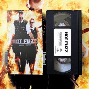 May include: A VHS tape and its cover for the movie "Hot Fuzz". The cover features two men in police uniforms with guns, set against a fiery background. The VHS tape is black with a white label that also says "Hot Fuzz".