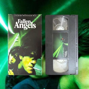 May include: A VHS tape and cover for the film "Fallen Angels" by Wong Kar-Wai. The cover has a green and black design with two people and the film title. The VHS tape is black with a label that says "Fallen Angels".