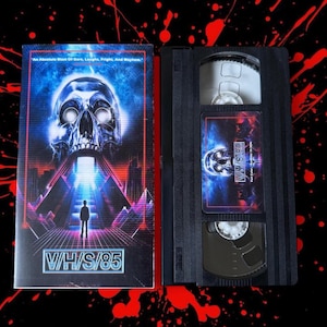 Peut inclure: Une cassette VHS et son boîtier avec le titre "V/H/S/85". Le boîtier présente un crâne aux yeux brillants au-dessus d'une silhouette et d'un paysage urbain futuriste. La cassette elle-même est noire avec une étiquette assortie.
