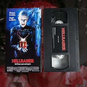 Könnte beinhalten: Ein Hellraiser VHS-Band und die dazugehörige Papphülle. Die Hülle zeigt eine bläuliche Illustration einer blassen Figur mit Nadeln im Kopf, die eine leuchtende Puzzle-Box hält, mit dem Text Demon to some. Angel to others. und He'll tear your soul apart.