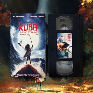 May include: A VHS tape and movie cover for the animated film "Kubo and the Two Strings." The cover features a silhouetted figure holding a sword against a stormy sea backdrop. The VHS tape is black with a label featuring a movie still.