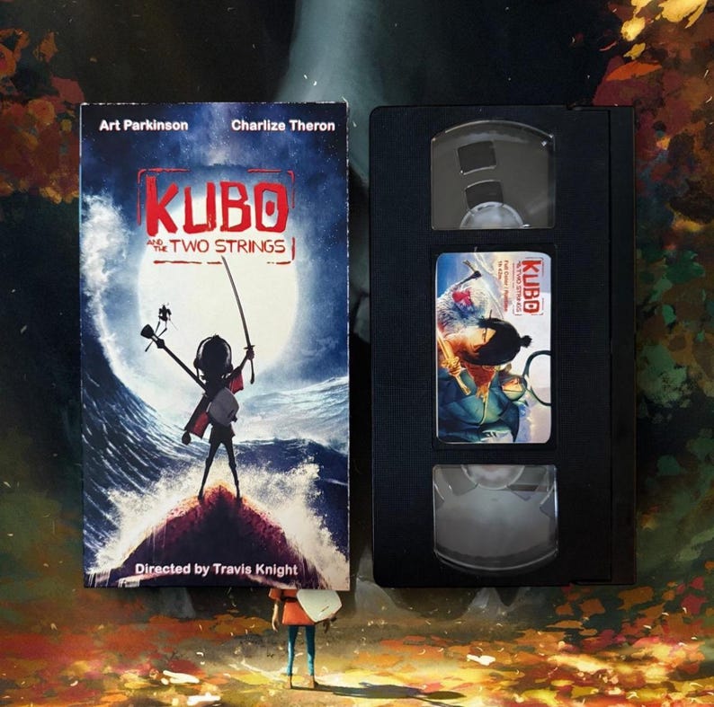 May include: A VHS tape and movie cover for the animated film "Kubo and the Two Strings." The cover features a silhouetted figure holding a sword against a stormy sea backdrop. The VHS tape is black with a label featuring a movie still.