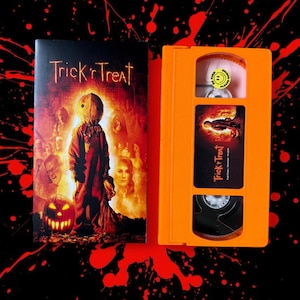Op de afbeelding: Een VHS-band en hoes voor de horrorfilm "Trick 'r Treat". De hoes toont een centrale figuur met een jute zakmasker, een pompoen en andere personages. De band is oranje met een bijpassend etiket.