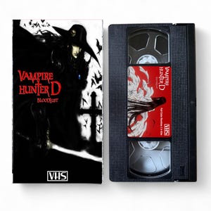 Puede incluir: Una cinta VHS y su estuche con el título "Vampire Hunter D: Bloodlust". El estuche tiene una ilustración oscura de un vampiro con el título en rojo y blanco. La cinta VHS es negra con una etiqueta roja.