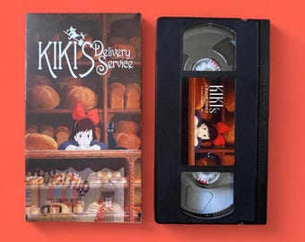 May include: A VHS tape and its cover for "Kiki's Delivery Service" are displayed against a red background. The cover features the film's title and a bakery scene with the main character. The VHS tape is black with a label showing the film's title and artwork.