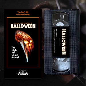Pode incluir: Uma fita VHS preta e sua capa para o filme "Halloween" de John Carpenter. A capa apresenta um design de faca e abóbora com o título do filme. A fita tem o título "Halloween" em texto branco.