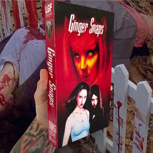 Puede incluir: Una cinta VHS de la pel&iacute;cula de terror "Ginger Snaps" con una portada roja que presenta a una mujer demon&iacute;aca y a otras dos mujeres. El t&iacute;tulo de la pel&iacute;cula tambi&eacute;n se muestra en el lomo de la cinta. La cinta se sostiene en una mano tatuada.