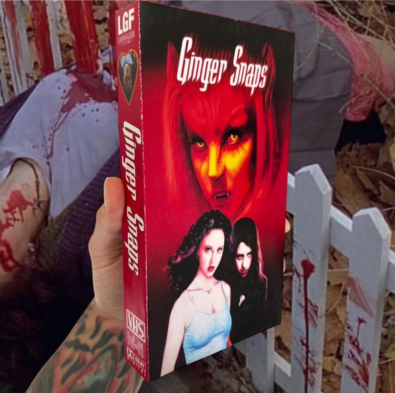 Puede incluir: Una cinta VHS de la pel&iacute;cula de terror "Ginger Snaps" con una portada roja que presenta a una mujer demon&iacute;aca y a otras dos mujeres. El t&iacute;tulo de la pel&iacute;cula tambi&eacute;n se muestra en el lomo de la cinta. La cinta se sostiene en una mano tatuada.