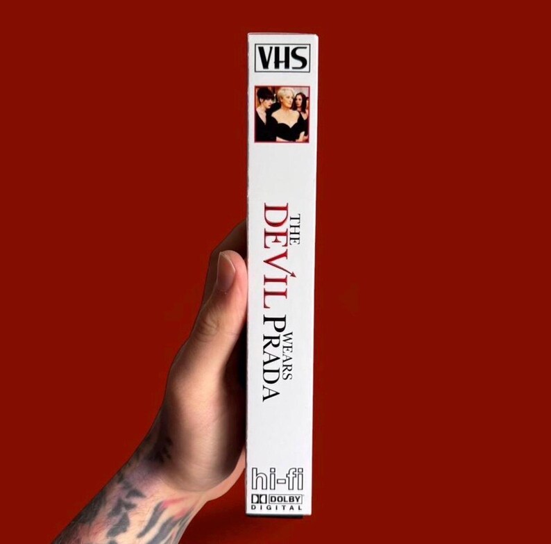 Puede incluir: Una cinta VHS blanca de la pel&iacute;cula "El diablo viste de Prada". La cinta tiene una peque&ntilde;a imagen del elenco en la parte superior y el t&iacute;tulo de la pel&iacute;cula en rojo y blanco. Tambi&eacute;n muestra los logotipos "hi-fi" y "Dolby Digital".