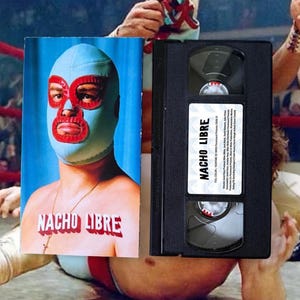 Może przedstawiać: Plakat filmowy i kaseta VHS do filmu "Nacho Libre". Plakat przedstawia luchadora w niebieskiej masce z czerwonymi detalami. Czarna kaseta VHS ma białą etykietę z napisem "Nacho Libre".