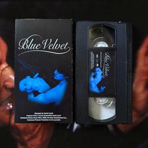 Puede incluir: Una cinta VHS y estuche vintage de la película "Blue Velvet". El estuche es negro con el título de la película en escritura blanca y una imagen azul. La cinta VHS también es negra, con el título de la película y una imagen azul.