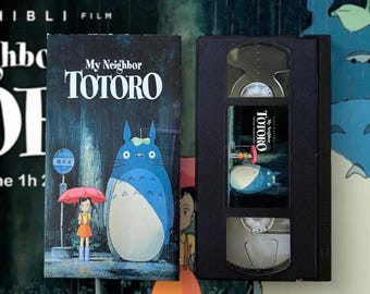 May include: A VHS tape and case for the animated film "My Neighbor Totoro." The case features a blue Totoro character with a girl holding an umbrella in the rain. The VHS tape is black with a label featuring the film's title and characters.