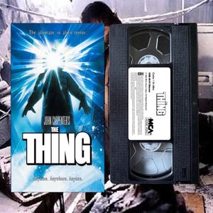 May include: A VHS tape and movie poster for John Carpenter's "The Thing." The poster features a figure with a bright light emanating from its head, with the movie title in large white letters. The VHS tape is black with a white label.