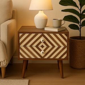 Puede incluir: Una mesita de noche de madera con dos cajones con un patrón de chevrón crema y marrón. Encima hay una lámpara blanca, una taza y un libro. La mesa mide aproximadamente 60 cm de alto.